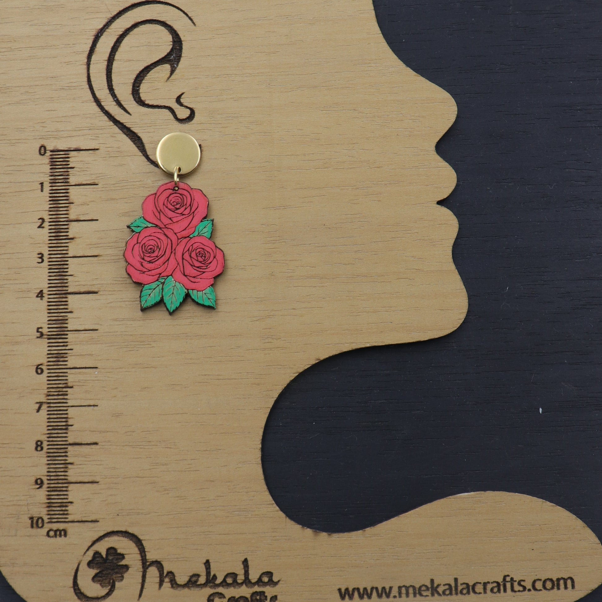 Earring with pink roses on a wooden scale next to a silhouette of a face, branded 'Mekala Crafts'.