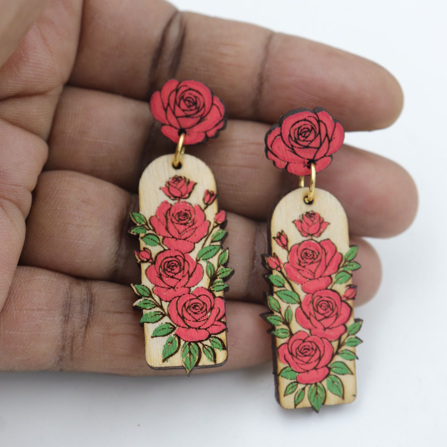 Wooden earrings with red rose designs held between fingers on a white background