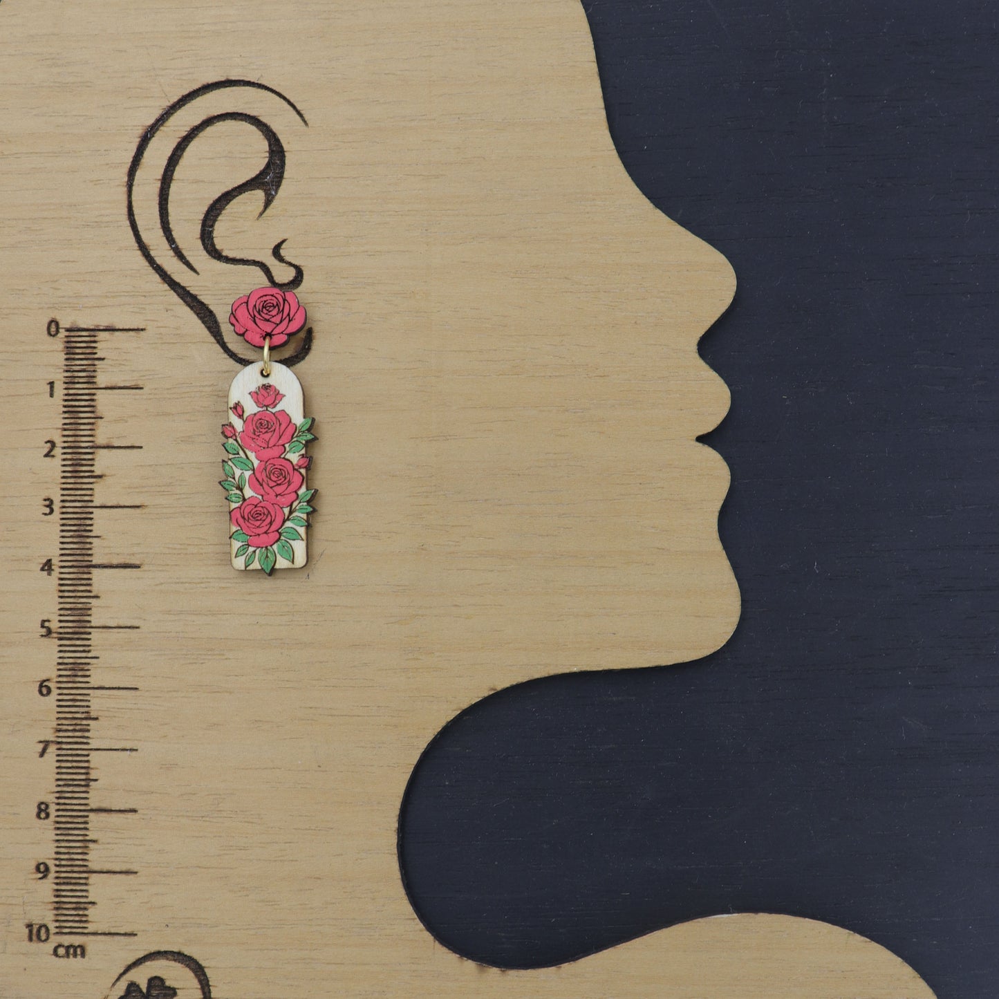 Floral earring displayed on a wooden block with an ear silhouette and ruler for scale.