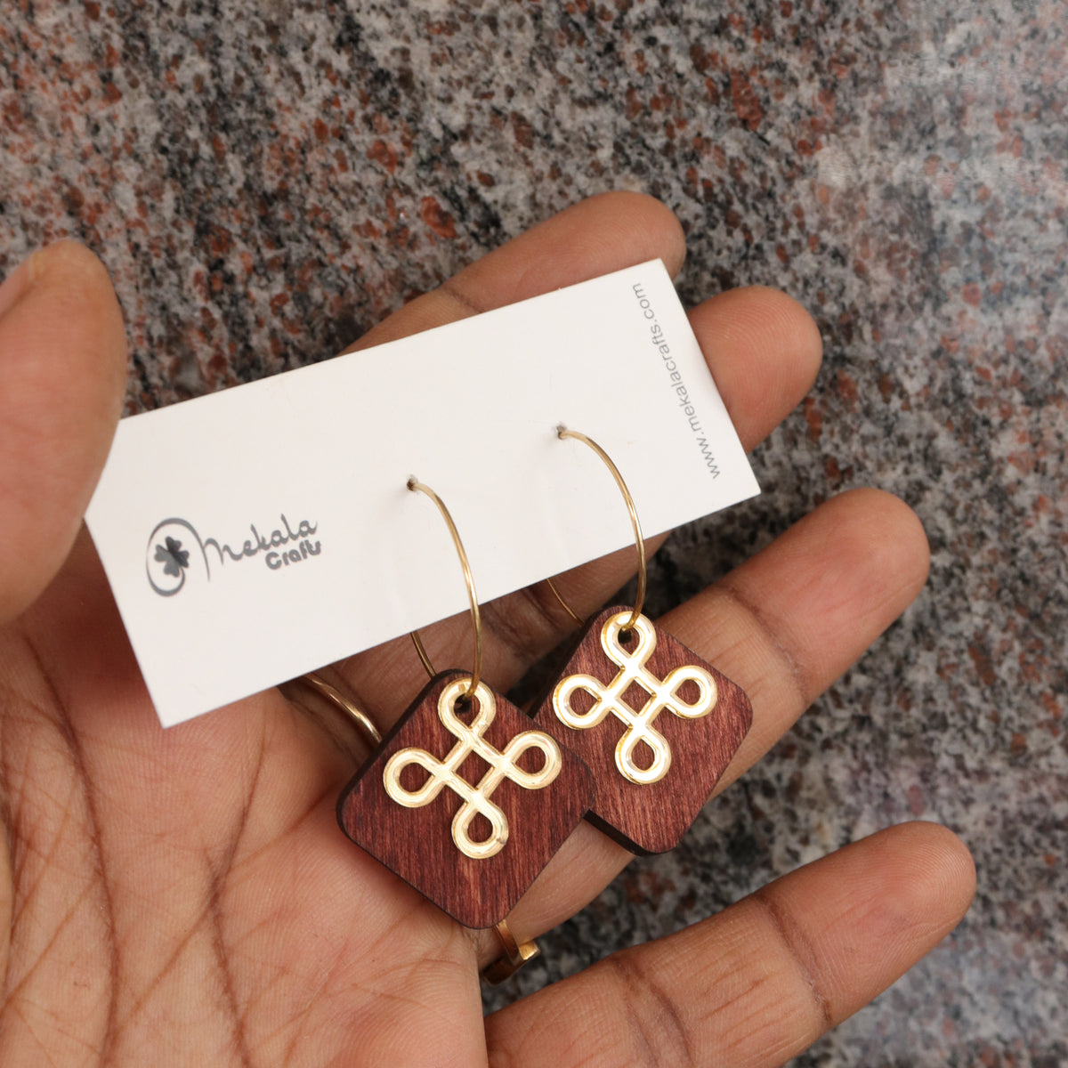 A pair of wooden drop earrings with gold Korean knot motifs, held in a hand over a speckled stone surface. The earrings are attached to a white display card with the brand name 'Mekala Crafts'.