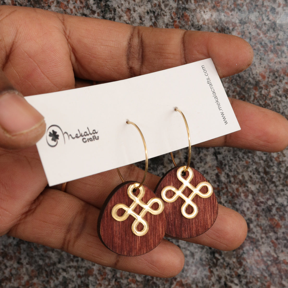 A close-up of a hand holding a white brand card displaying a pair of wooden dangle earrings with gold Celtic knot designs on a speckled gray countertop.